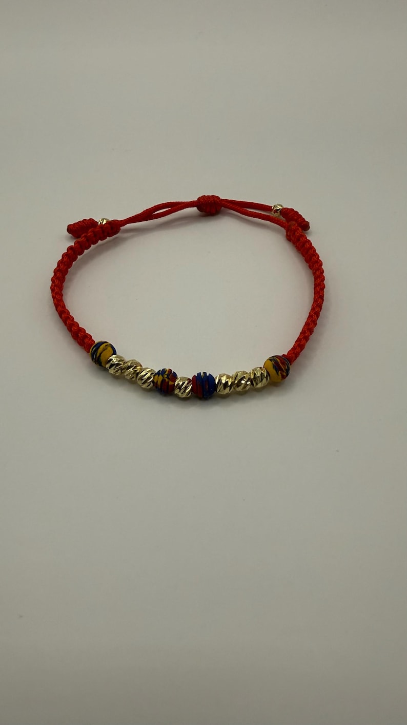 May include: Red string bracelet with gold-coloured beads and colourful accent beads. The bracelet has a sliding knot closure for adjustable sizing. The beads have a textured, ridged design.