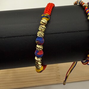 May include: Two bracelets are shown. One has gold-coloured beads and red, blue, and yellow accents. The other bracelet is partially visible. The bracelets are displayed on a black surface.