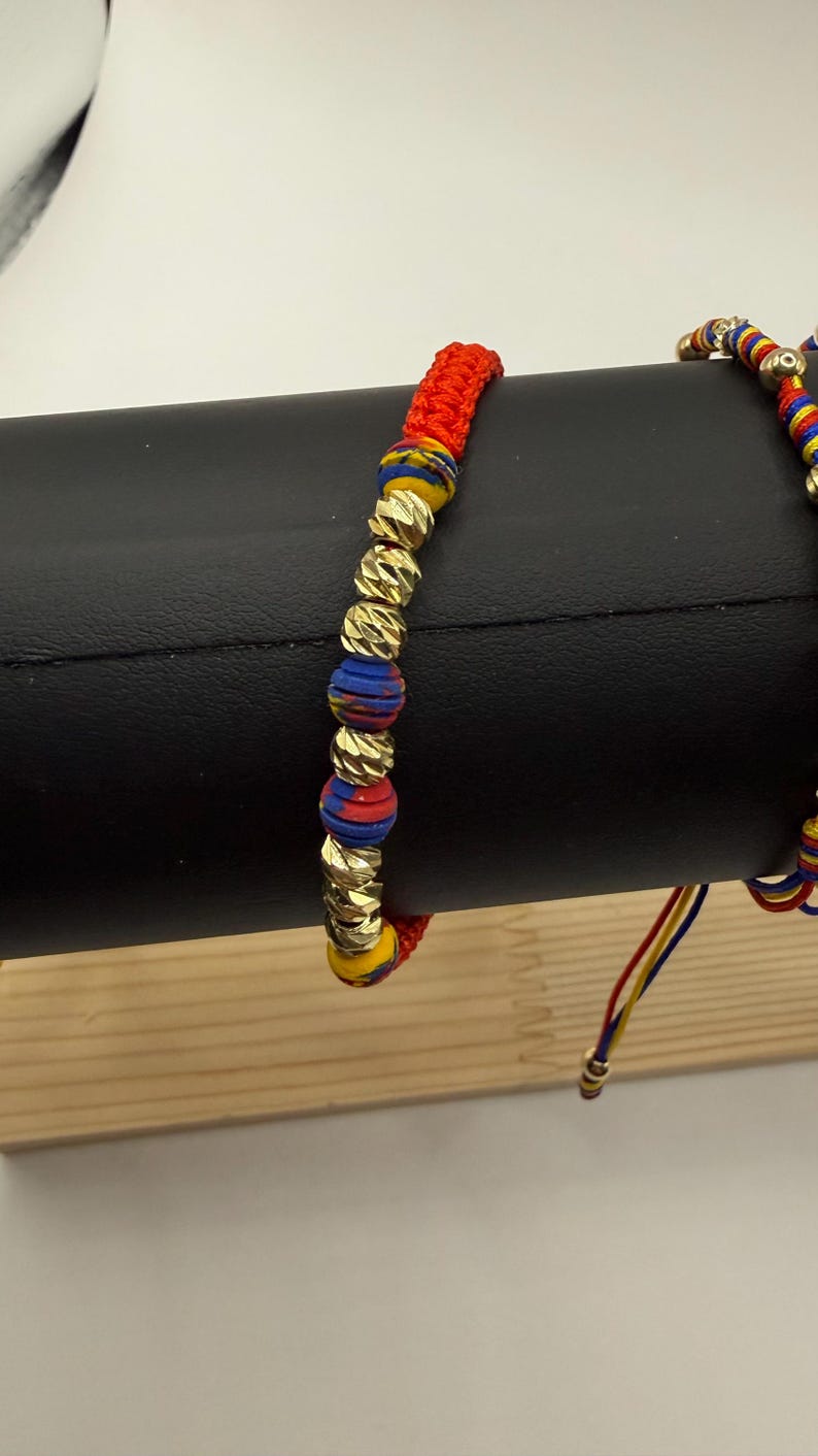 May include: Two bracelets are shown. One has gold-coloured beads and red, blue, and yellow accents. The other bracelet is partially visible. The bracelets are displayed on a black surface.