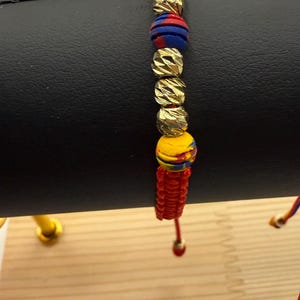 May include: A colourful bracelet with gold-coloured beads and vibrant red, blue, and yellow accents. The bracelet is strung on a red cord and has a unique design. The beads have a textured surface.