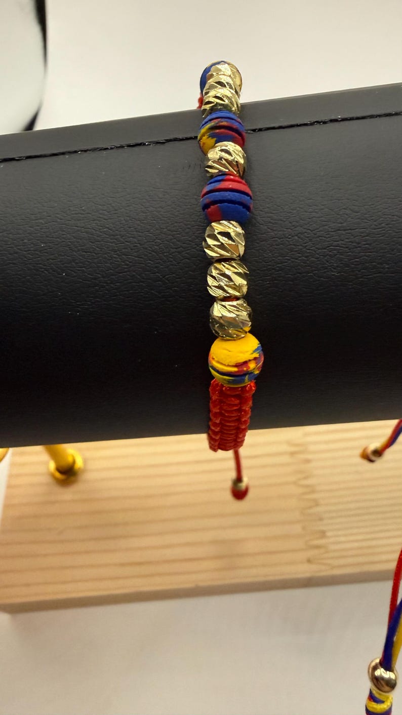 May include: A colourful bracelet with gold-coloured beads and vibrant red, blue, and yellow accents. The bracelet is strung on a red cord and has a unique design. The beads have a textured surface.