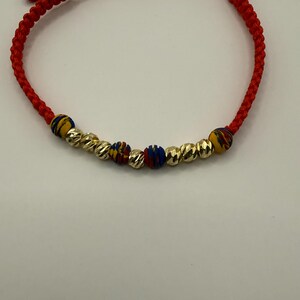 May include: Red braided bracelet featuring gold-coloured and patterned beads. The bracelet has an adjustable sliding knot closure. The gold beads have a textured, ridged design.