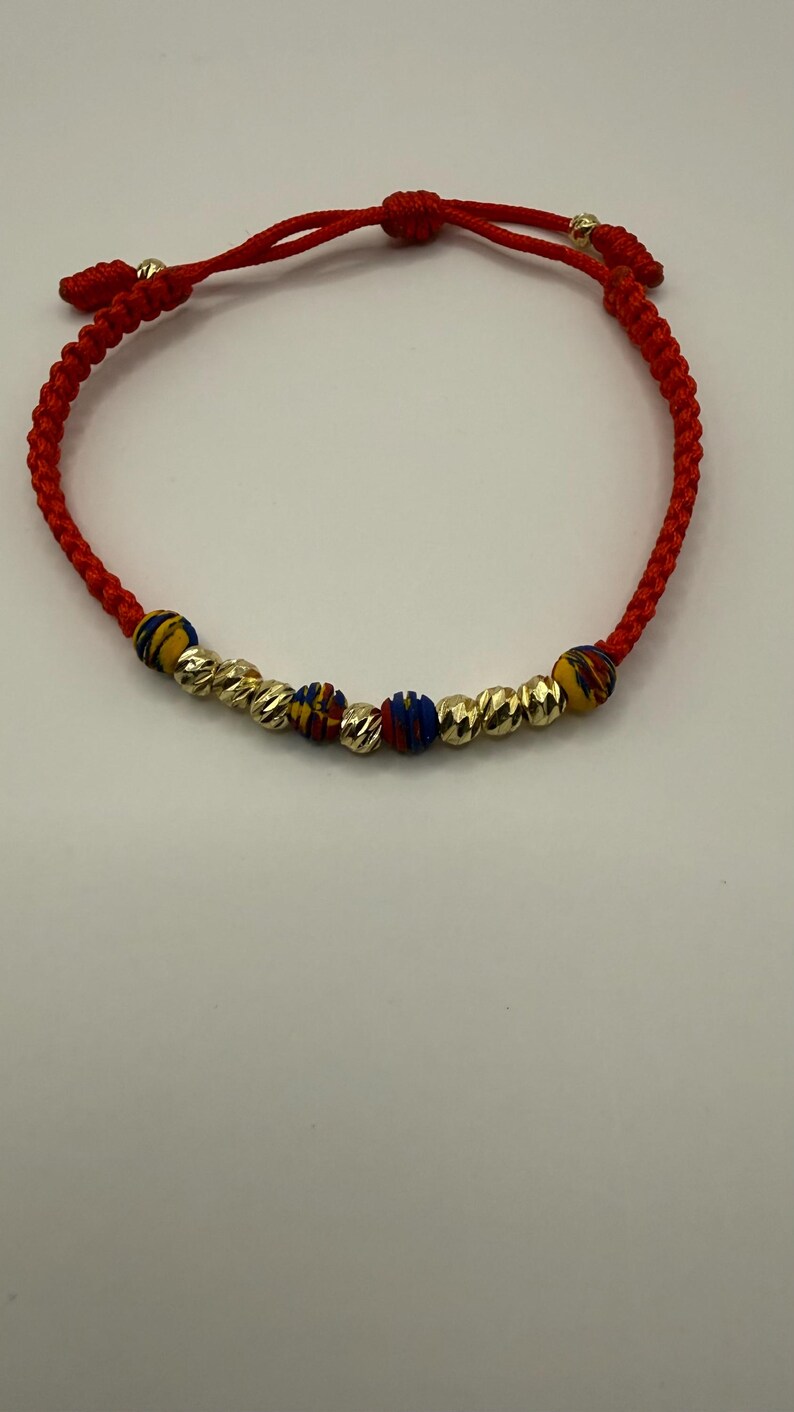 May include: Red braided bracelet featuring gold-coloured and patterned beads. The bracelet has an adjustable sliding knot closure. The gold beads have a textured, ridged design.