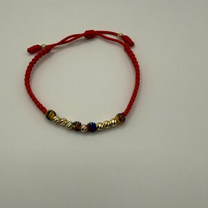 May include: Red braided bracelet with gold-coloured beads and accents. The bracelet features textured gold beads and smaller beads in blue, yellow, and red. The bracelet is adjustable.
