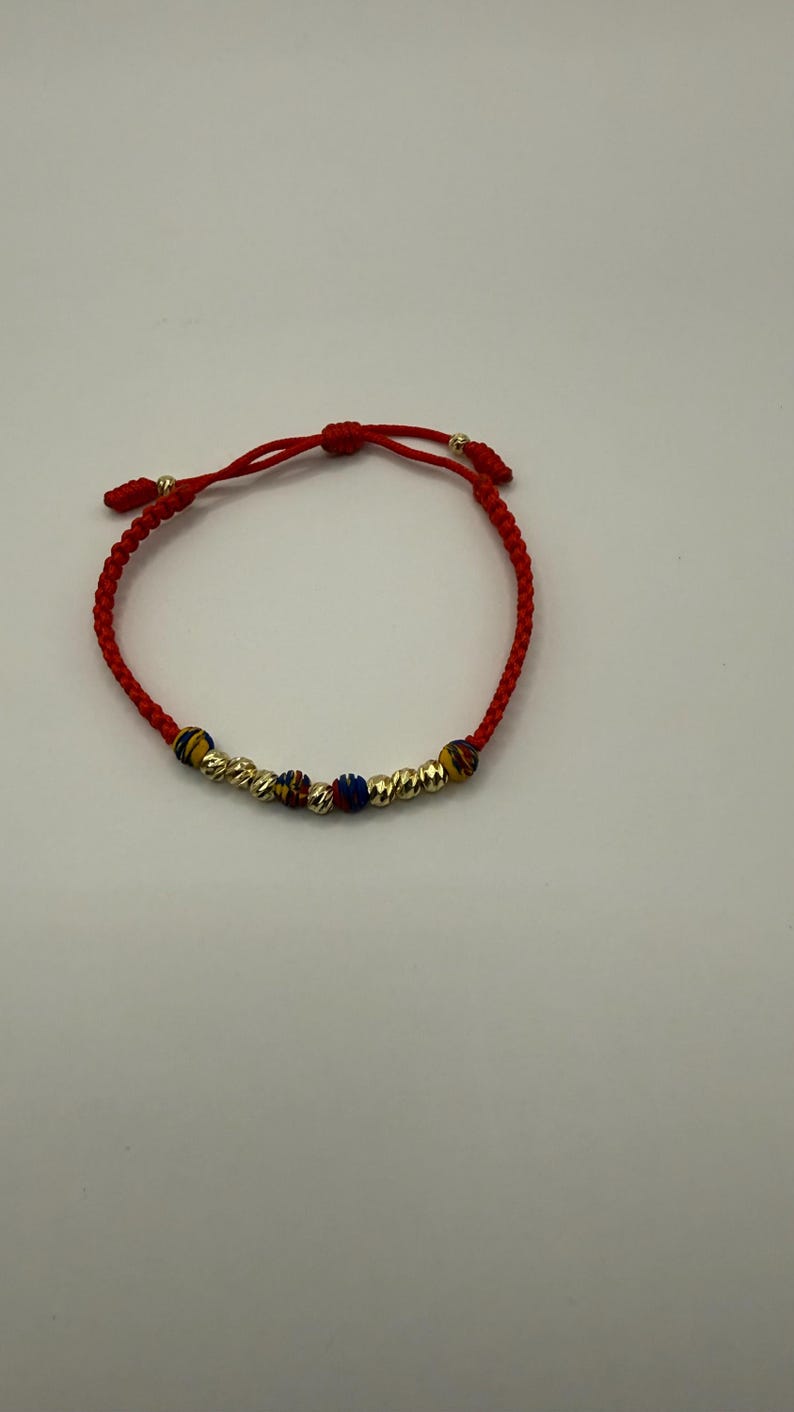 May include: Red braided bracelet with gold-coloured beads and accents. The bracelet features textured gold beads and smaller beads in blue, yellow, and red. The bracelet is adjustable.