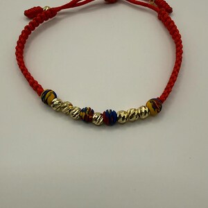 May include: Red braided bracelet with gold-coloured beads and colourful accent beads. The bracelet has a sliding knot closure for adjustable sizing. The gold beads have a textured surface. A simple and elegant piece of jewellery.
