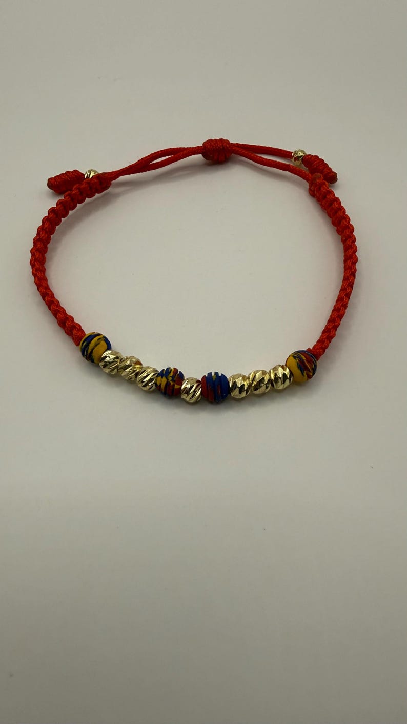 May include: Red braided bracelet with gold-coloured beads and colourful accent beads. The bracelet has a sliding knot closure for adjustable sizing. The gold beads have a textured surface. A simple and elegant piece of jewellery.