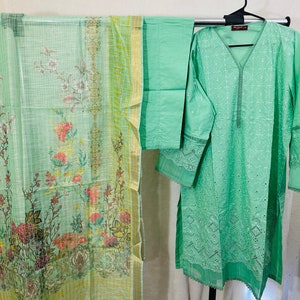 May include: A green embroidered dress with a matching dupatta and pants. The dress has a V-neckline and long sleeves with lace detailing. The dupatta has a floral print and the pants are plain.