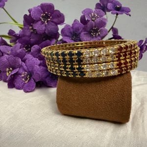 May include: Gold bangles with alternating sections of navy blue, white, and red stones. The bangles are stacked on a brown velvet stand, with purple flowers in the background. The jewelry is displayed on a white cloth.