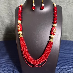 May include: A red beaded necklace with gold accents and matching earrings. The necklace features a layered design with multiple strands of small red beads and two larger red beads with gold accents. The earrings are simple and feature a single red bead with a gold accent.