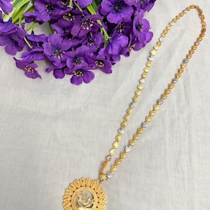 May include: A gold-toned necklace with a large, round pendant featuring a detailed design. The necklace chain alternates between gold and silver-toned heart-shaped links. Purple flowers are in the background.