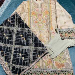 May include: A beige embroidered dress with a black and gold net dupatta. The dress has a floral design and the dupatta has a geometric pattern. The dress is trimmed with gold beads.