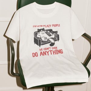 Könnte beinhalten: Weißes T-Shirt mit einer Grafik einer Katze auf einer Couch, die eine Fernbedienung hält. Der Text auf dem Shirt lautet: "STOP HATING ON LAZY PEOPLE WE DIDN'T EVEN DO ANYTHING". Das Shirt liegt auf einem grünen Bürostuhl.