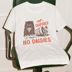 Può includere: T-shirt bianca con grafica di un gatto soffice, un vaso di fiori rovesciato e la scritta "all + oopsies, no daisies". La maglietta è appoggiata su una sedia da ufficio verde con struttura in metallo.