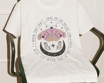 May include: White t-shirt with a graphic of a pink and purple butterfly with a crescent moon and stars. The text reads "When you can't look on the bright side, I will sit with you in the dark."