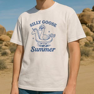 Può includere: T-shirt bianca con grafica blu di un'oca con cappello e un unicorno su un galleggiante gonfiabile. Il testo "SILLY GOOSE Summer" è stampato in blu sopra e sotto la grafica. La maglietta è realizzata in materiale morbido.