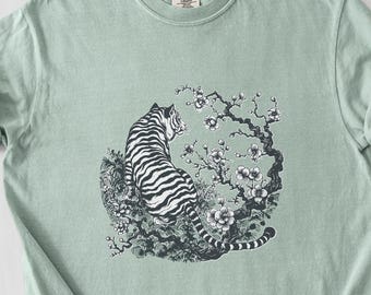 以下が含まれることがあります： 白い花が咲いた木に腰掛けた黒と白の虎のイラストが描かれた薄い緑色のTシャツ。虎は背を向けています。