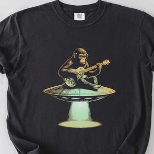 May include: Black t-shirt with a graphic of a monkey playing a guitar on a flying saucer. The monkey is wearing a red bandana and the guitar is yellow.