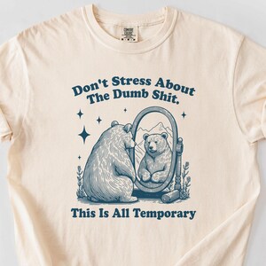 May include: A cream colored t-shirt with a blue graphic of a bear looking in a mirror. The text on the shirt reads "Don't Stress About The Dumb Shit. This Is All Temporary."