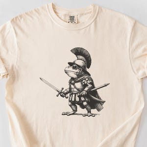 Op de afbeelding: Een crèmekleurig T-shirt met een zwart-witte illustratie van een kikker die een helm draagt en een zwaard vasthoudt.