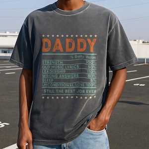 May include: A dark grey t-shirt with a vintage look featuring the word "DADDY" in orange and a list of qualities with percentages. The list includes "Strength", "Old Music Lyrics", "Leadership", "Wrong Answers", "Sleep", "Unconditional Love", and "Still the Best Job Ever".