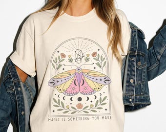 May include: A cream colored t-shirt with a graphic of a moth with a crystal and floral accents. The text "MAGIC IS SOMETHING YOU MAKE" is printed below the graphic.