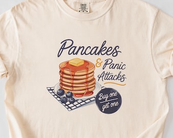 Puede incluir: Una camiseta beige con un gráfico de una pila de panqueques con arándanos y el texto "Pancakes & Panic Attacks" y "Buy one get one".