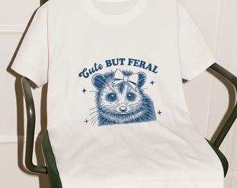 May include: White t-shirt with a blue illustration of an opossum wearing a bow and the text "Cute But Feral".