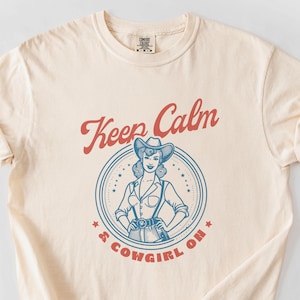 Può includere: Una t-shirt color crema con una grafica in stile vintage che dice "Keep Calm & Cowgirl On". La grafica presenta una cowgirl dei cartoni animati con un cappello da cowboy e un vestito blu e bianco.