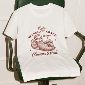 Puede incluir: Camiseta blanca con una ilustración de un perezoso marrón y el texto "Relax We're All Crazy it's not a Competition".