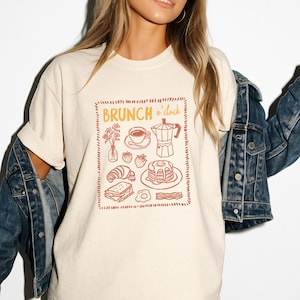 May include: Cream-colored t-shirt featuring a brunch-themed graphic. The design includes a coffee cup, strawberries, pancakes, and a croissant, with the words "BRUNCH o'clock" in orange. The shirt is made of a soft material.
