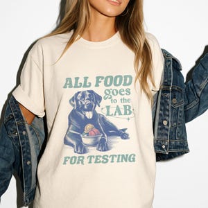 Puede incluir: Una camiseta blanca con un labrador retriever negro sosteniendo un tazón de helado. El texto en la camiseta dice "ALL FOOD goes to the LAB FOR TESTING".