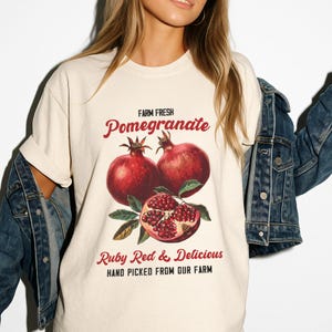 May include: Cream-colored t-shirt featuring a vintage-style illustration of pomegranates. The design includes the words "Farm Fresh Pomegranate" and "Ruby Red & Delicious, Hand Picked From Our Farm" in a red script font. The shirt is worn by a person.
