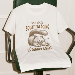 May include: White t-shirt with a brown illustration of a cat and the text "The Only Squat I'm Doing Is Diddly Squat."