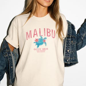 May include: Cream-colored t-shirt with the word "MALIBU" in pink, above a blue turtle graphic with pink flowers. Below the graphic, the text reads "CATCH WAVE SURF MEMORIES SIESTA BEACH". The shirt has a relaxed fit.