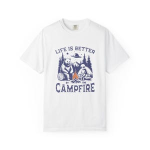 Puede incluir: Camiseta blanca con un diseño gráfico azul marino. El diseño muestra un oso, un extraterrestre y una escena de fogata. El texto "LIFE IS BETTER BY THE CAMPFIRE" está impreso arriba y abajo.