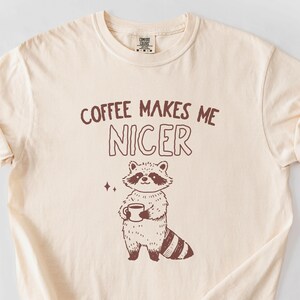 May include: A beige t-shirt with a brown raccoon illustration holding a cup of coffee. The text "Coffee Makes Me Nicer" is printed on the shirt.