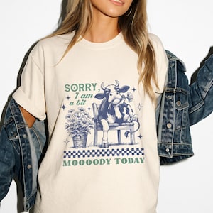 May include: A white t-shirt featuring a blue and white illustration of a cow sitting on a bench with a flower in its mouth. The text "SORRY, I am a bit MOOOODY TODAY" is printed on the shirt.