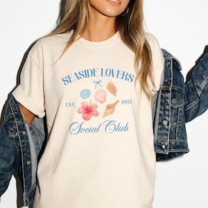 May include: Cream-colored t-shirt with the text "Seaside Lovers Social Club" in light blue script. The design includes watercolor illustrations of seashells, a hibiscus flower, and the year 1997. The shirt is paired with a denim jacket.