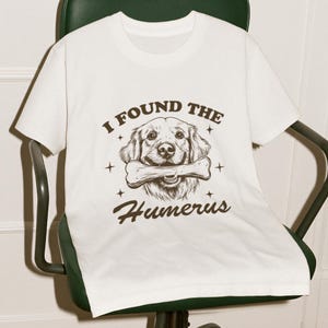 May include: White t-shirt with a brown graphic of a golden retriever dog holding a bone in its mouth. The text "I found the humerus" is printed below the dog.