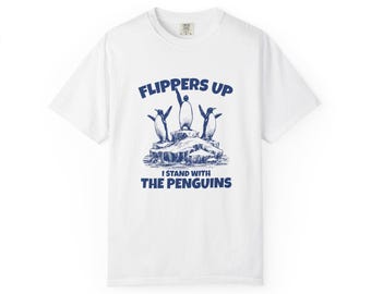 May include: White t-shirt with a blue graphic of three penguins on an iceberg, with the text "FLIPPERS UP" above and "I STAND WITH THE PENGUINS" below. The shirt has a crew neck and short sleeves.