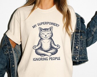 May include: Cream-colored t-shirt featuring a navy blue illustration of a cat in a yoga pose. The text above the cat reads "MY SUPERPOWER?" and below it, "IGNORING PEOPLE". The shirt has short sleeves and a crew neck.