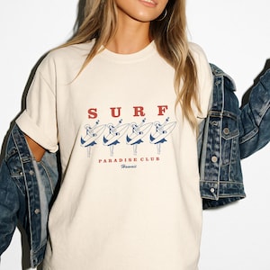 May include: Cream-colored t-shirt with the word "SURF" in red letters above a graphic of four surfers in blue and white. Below the graphic, the text reads "PARADISE CLUB Hawaii". The shirt has a relaxed fit.