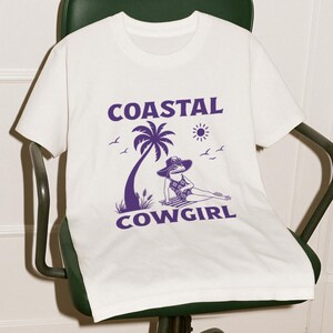 May include: White t-shirt with a purple graphic. The graphic features the text "COASTAL COWGIRL" with a palm tree, sun, and a frog wearing a hat and swimsuit. The frog is lounging on a beach towel.
