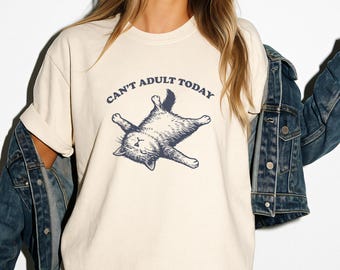 May include: Cream-colored t-shirt with the text "Can't Adult Today" above a navy blue illustration of a relaxed cat. The shirt is made of a soft material and has a classic crew neck design. The model is wearing a denim jacket.