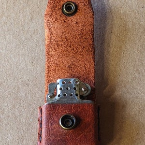 May include: A brown leather lighter case with a metal lighter partially visible. The case has a snap closure and a loop at the top. The leather has a textured appearance.