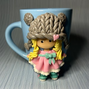 Puede incluir: Una taza de cerámica azul claro con un diseño de muñeca de arcilla hecha a mano. La muñeca lleva un gorro de punto gris, un vestido rosa y tiene el pelo rubio.