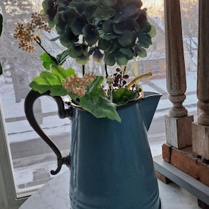 May include: A blue enamel pitcher with a silver handle filled with a bouquet of green and white flowers.