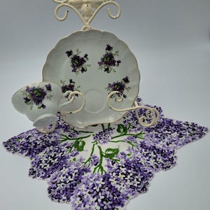 May include: A white teacup and saucer set with purple floral designs, displayed on a decorative metal stand. The set rests on a purple and white floral patterned doily. The stand is cream-colored.