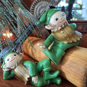 May include: Two ceramic elf figurines wearing green hats and outfits with gold-colored vests. One elf is reclining on its side, while the other is positioned in a seated pose. Both figures feature painted facial details, including red smiles and dark eyes.